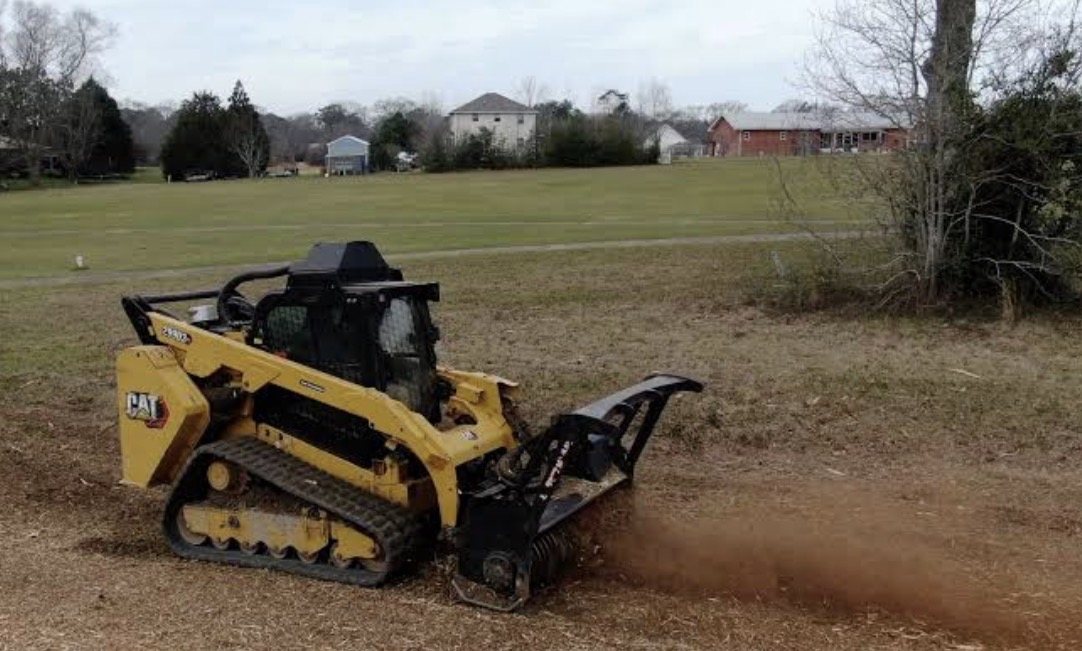 Skid steer mulching an open field with houses in the distance